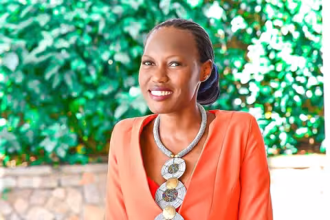 Brendah is wearing an orange jacket with a patterned necklace stands against a leafy green background.