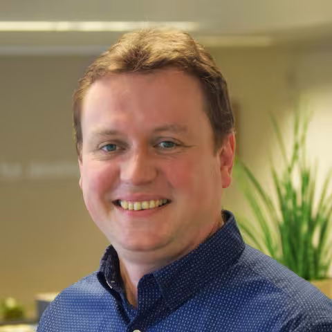 Koenraad is smiling while wearing a blue shirt. The background is a blurred office.