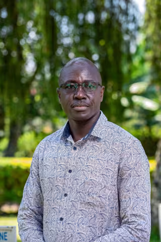 Louis is wearing glasses and a paisley-patterned shirt stands outdoors, looking serious. The background is lush with greenery.