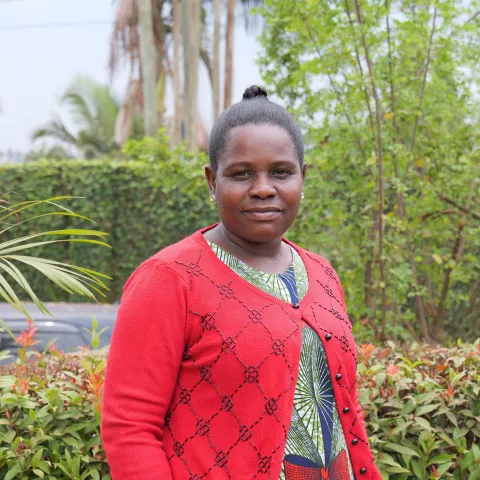 Lydia stands in a garden, wearing a red cardigan with a patterned dress. She smiles softly, surrounded by lush greenery and tall trees.