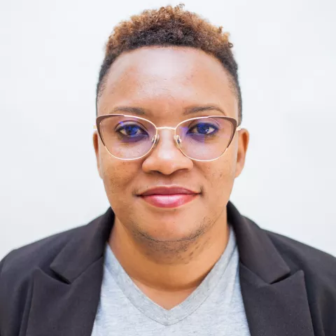 Maimbolwa is wearing glasses and smiles warmly. She is wear a grey shirt and a black blazer, conveying a friendly, professional look. The background is plain white.