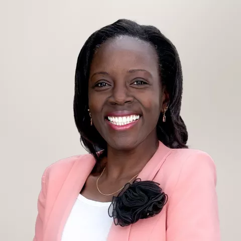 Mary is wearing a pink blazer with a black flower accessory on the lapel. The background is light gray.