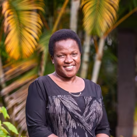 Milly is wearing black patterned dress and stands outdoors with lush green and yellow palm leaves in the background.