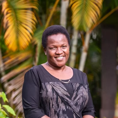Milly is wearing black patterned dress and stands outdoors with lush green and yellow palm leaves in the background.