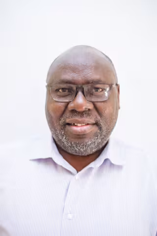 Musonda is wearing a light-striped shirt and glasses against a plain white background