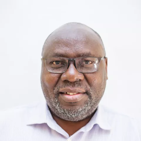 Musonda is wearing a light-striped shirt and glasses against a plain white background