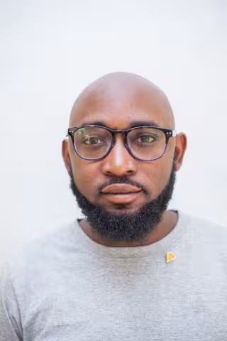 Mwango is wearing black glasses and grey t-shirt. The background is plain white.