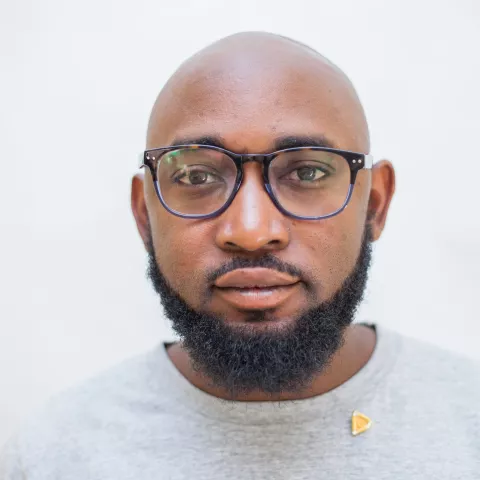 Mwango is wearing black glasses and grey t-shirt. The background is plain white.