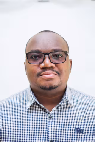 Mwanza is wearing a checkered shirt and glasses. The background is plain white.