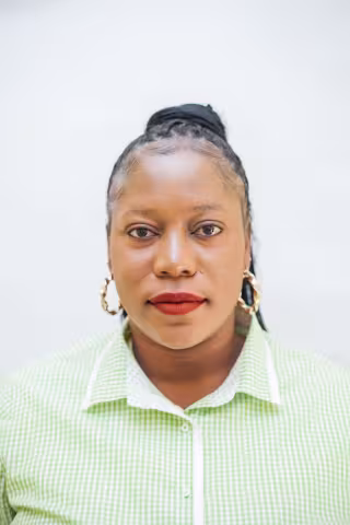 Nancy is wearing a white and green checkered shirt and gold hoop earrings. She looks directly at the camera. The background is plain white.