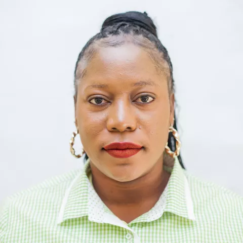 Nancy is wearing a white and green checkered shirt and gold hoop earrings. She looks directly at the camera. The background is plain white.