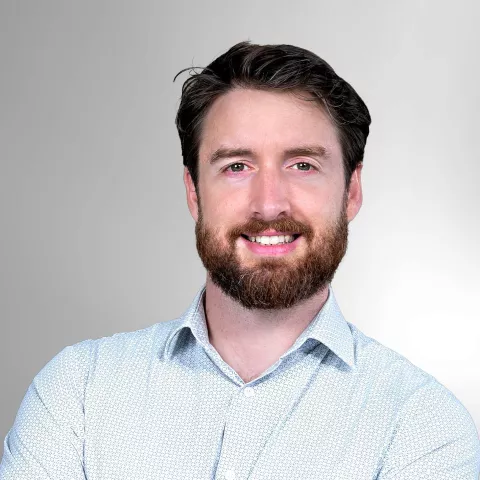 Noam smiles warmly, wearing a light blue patterned shirt. He stands against a light gray background.