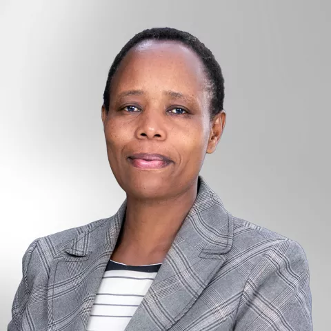 Peninah is wearing a gray plaid blazer and striped shirt smiles subtly against a light gray background.