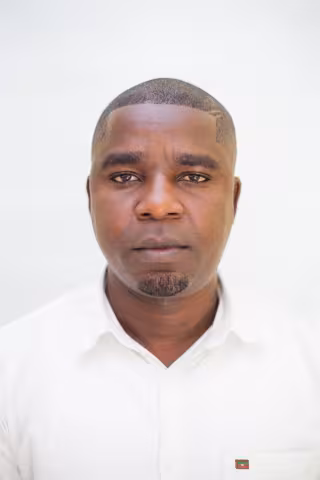 Prosper is wearing a white shirt as he looks directly into the camera. The background is plain white.
