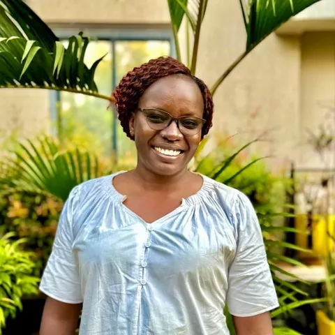 Rita is wearing glasses and shiny blouse. She stands outdoors with lush green plants and a beige building in the background