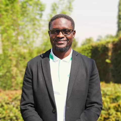 Robert is wearing glasses, a white shirt, and a black blazer. Background is sunlit greenery.