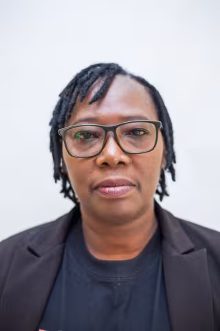 Sandra has a neutral expression. She wears glasses, a black blazer and black shirt. The background is plain white. 