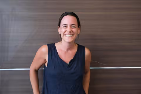 Silvija is wearing a blue top. She is smiling and standing in front of a brown wall. 