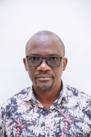Siza is wearing glasses and a floral shirt. He stands against a plain white background, looking directly at the camera with a neutral expression.