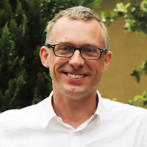 Stefaan is wearing a white shirt and glasses. The background is lush green plants.