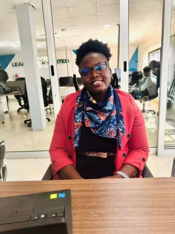 Theresa is wearing glasses, a red blazer, and a floral scarf. She smiles while sitting in a modern office with glass dividers and computer workstations.