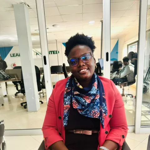 Theresa is wearing glasses, a red blazer, and a floral scarf. She smiles while sitting in a modern office with glass dividers and computer workstations.