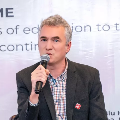 Tom is speaking into a microphone. He is wearing a grey blazer over a checkered shirt. 