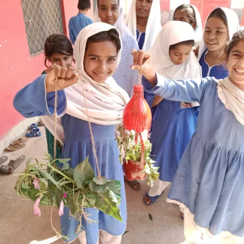 Smiling girls in blue and white uniforms hold hanging plant crafts made from recycled bottles. They stand outside a red wall, exuding happiness and creativity.