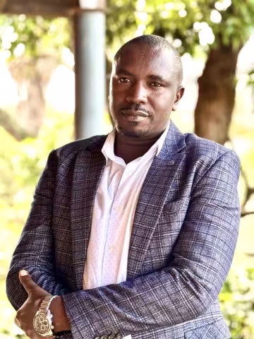 Male staff member stands outdoors with arms loosely crossed, wearing a checked blazer over a light-coloured button-up shirt with trees and greenery in the background.