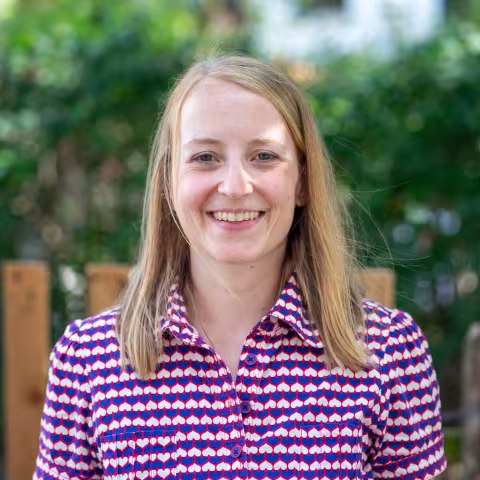 A smiling woman with straight, shoulder-length blonde hair stands outdoors in front of a softly blurred background of green foliage and a wooden fence. She is wearing a short-sleeved collared shirt with a red, white and blue geometric pattern.