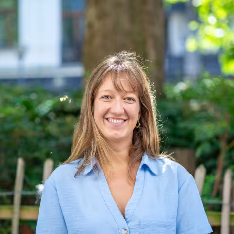 A smiling woman with shoulder-length light brown hair stands outdoors wearing a light blue blouse, with a wooden fence and leafy trees softly blurred in the background.
