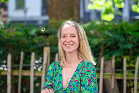 Claire is smiling. She has shoulder-length blonde hair stands outdoors in a green floral dress, with a wooden fence and leafy trees in the background.
